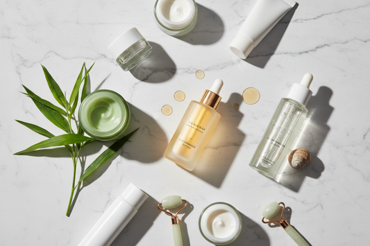 Editorial flat-lay of K-beauty skincare products on white marble — niacinamide serum, centella asiatica cica cream with tiger grass leaves, and snail mucin essence — the three most popular Korean beauty ingredients for glowing, healthy skin