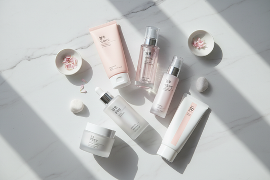Flat lay of Korean beauty morning skincare routine products including cleanser, toner, essence, serum, moisturizer, and sunscreen arranged on a white marble surface