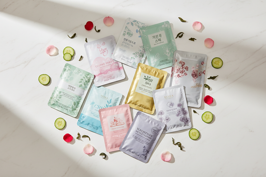 "Flat lay of colorful Korean sheet masks arranged on a white surface with fresh cucumber slices, rose petals, and green tea leaves representing different K-Beauty mask ingredients"